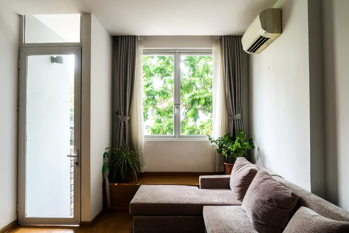 A modern living room with a gray sectional sofa, potted plants, a window with curtains, a glass door, and a wall-mounted ductless HVAC air conditioner.