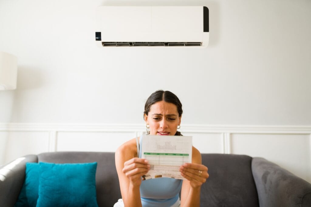 Reduce AC Costs This Summer With These HVAC Tips 1 A woman sits on a couch under an air conditioner, looking concerned as she reads a bill or statement, wondering how to reduce AC costs.