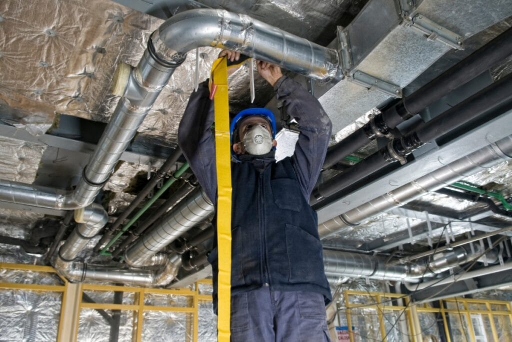 Reduce AC Costs This Summer With These HVAC Tips 2 A worker wearing a hard hat and face mask insulates ductwork on a ceiling in an industrial building under construction to help reduce AC costs.