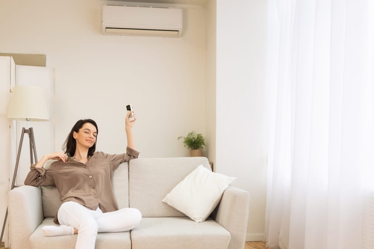 Woman sitting on a sofa using a remote control to operate an air conditioner in a bright living room, looking for ways to reduce AC costs.