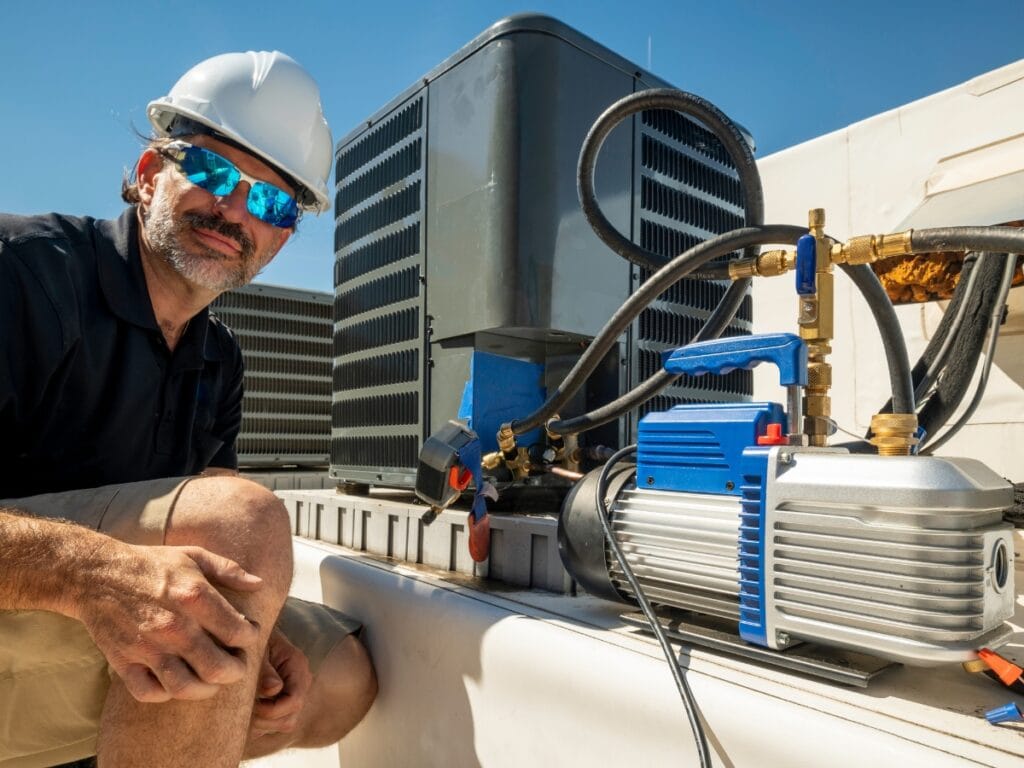 Commercial HVAC Maintenance Tips for Fall 11 A technician wearing a hard hat and sunglasses kneels next to rooftop commercial HVAC units and equipment under a clear blue sky, ready to perform essential Commercial HVAC maintenance.