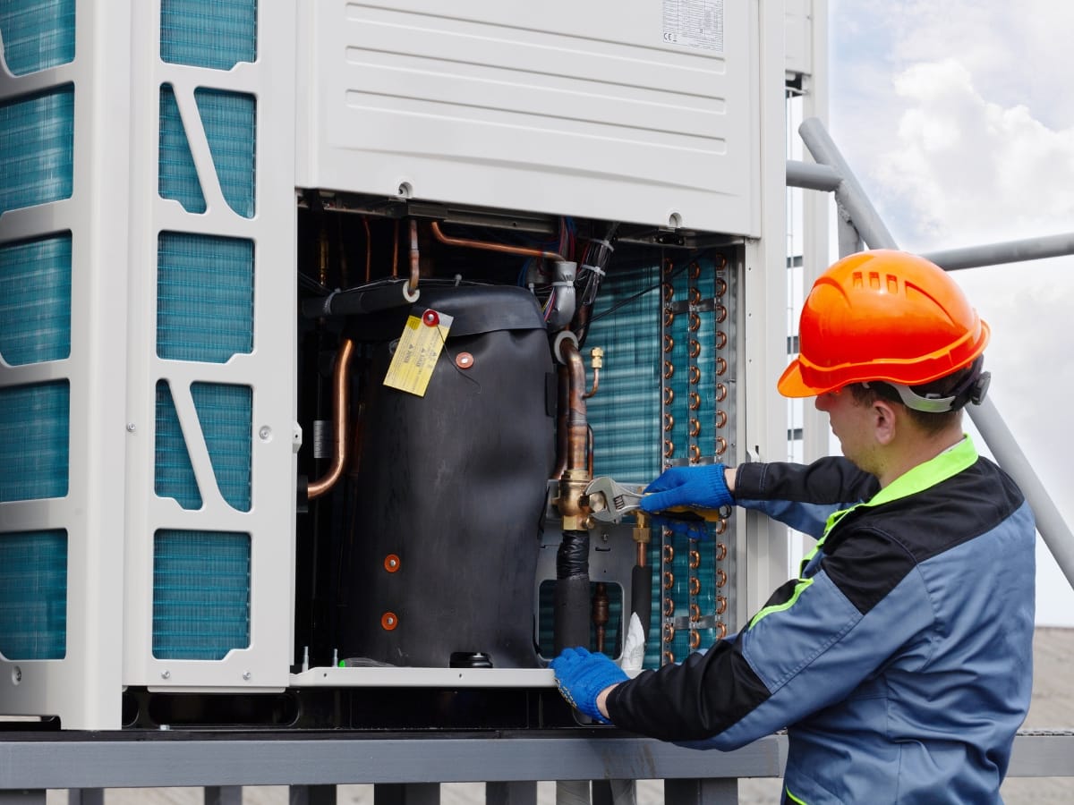 A technician wearing safety gear inspects and adjusts pipes inside an outdoor commercial HVAC unit, demonstrating the importance of regular Commercial HVAC maintenance and fall HVAC tips for optimal performance.