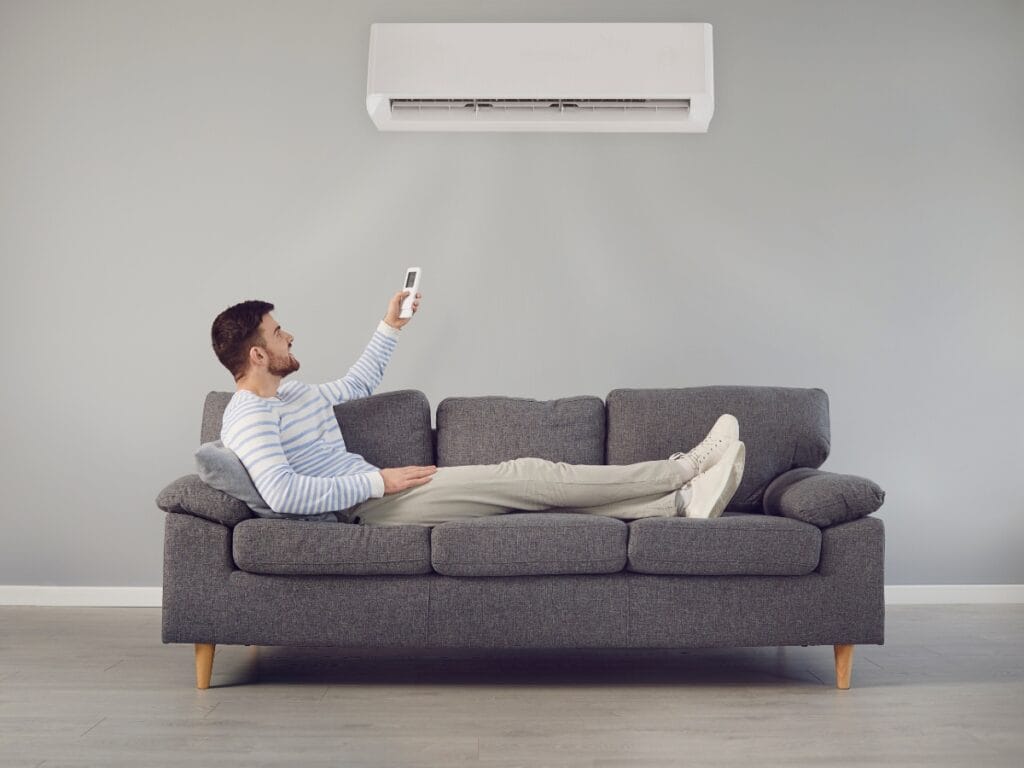 A man lying on a gray sofa uses a remote control to operate an air conditioner mounted on a light gray wall, ensuring comfort and following his HVAC Checklist for optimal performance.
