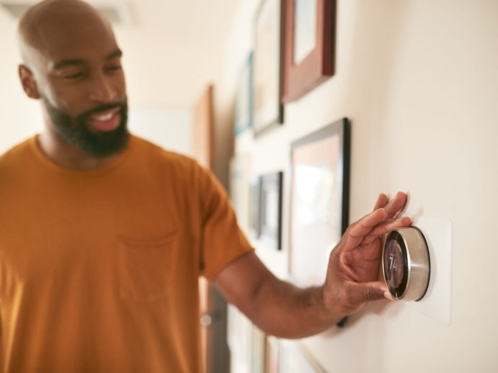 A man in an orange shirt adjusts a smart thermostat on a wall in a hallway decorated with framed pictures, following his HVAC checklist for optimal home comfort.
