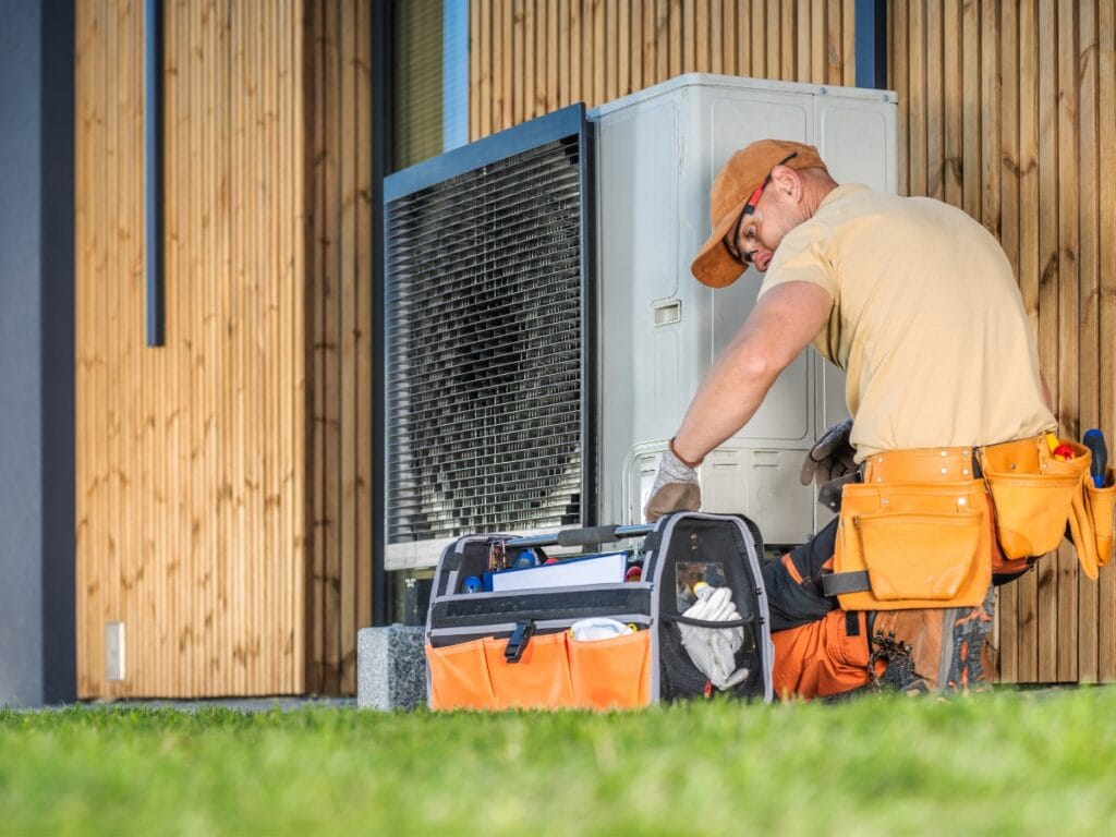 A technician wearing an orange cap and tool belt follows an HVAC checklist while repairing an outdoor air conditioning unit next to a building with wooden siding. A tool bag rests on the grass nearby.