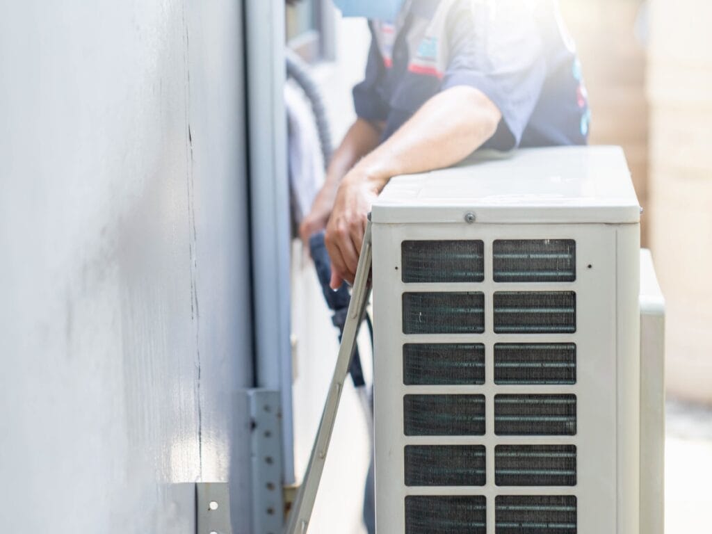 A technician, wearing a blue uniform, performs annual HVAC maintenance while installing or repairing an outdoor air conditioning unit next to a building wall.