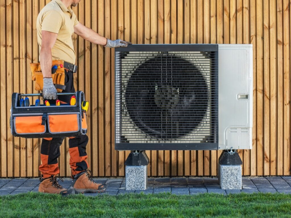 A technician wearing gloves and work clothes stands beside an outdoor HVAC unit, holding an orange and blue tool bag, ready to perform annual HVAC maintenance.