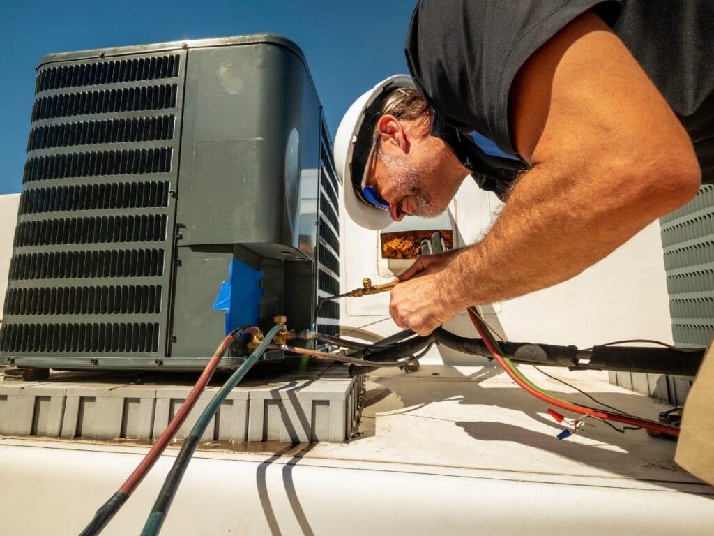 A technician in a hard hat and safety glasses performs annual HVAC maintenance, repairing an outdoor air conditioning unit on a rooftop using various tools and gauges.