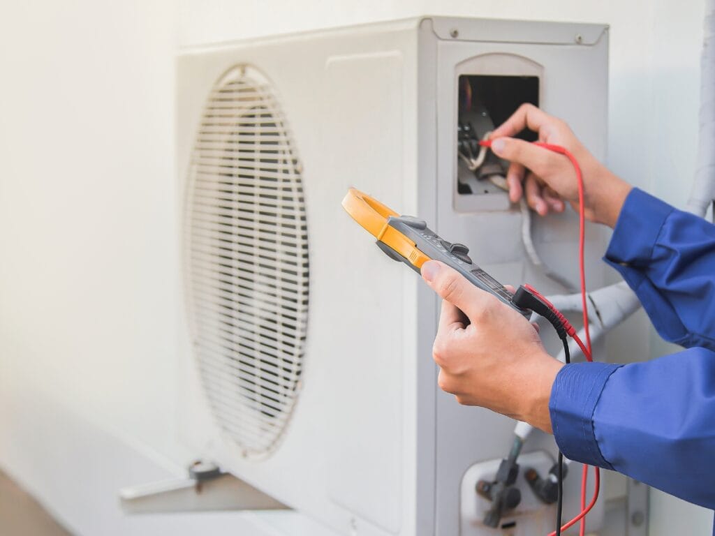 A person in blue coveralls uses a digital multimeter to check the electrical connections of an outdoor air conditioning unit, addressing common HVAC replacement questions during the inspection.