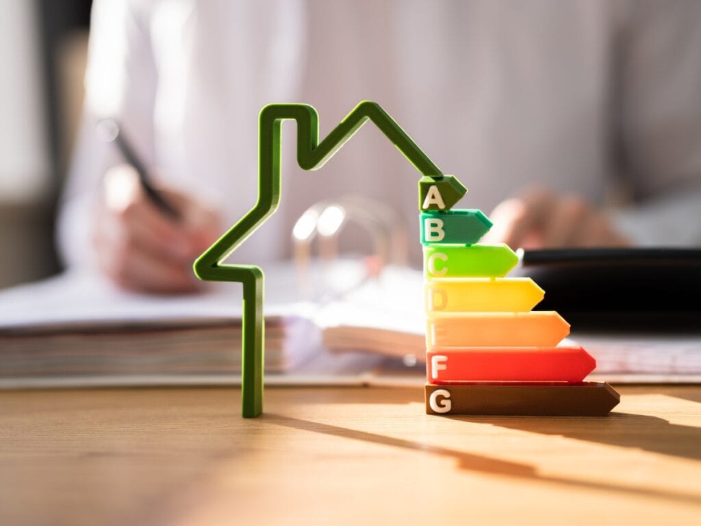 A person sits at a desk with a pen and papers, behind a model of a house and a colorful energy efficiency rating scale from A to G, carefully considering HVAC Replacement Questions.