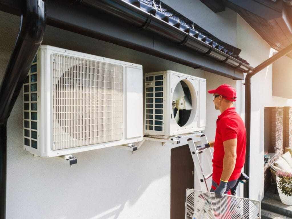 A technician in a red uniform stands on a ladder inspecting two outdoor air conditioning units mounted on a building wall, addressing common HVAC replacement questions.