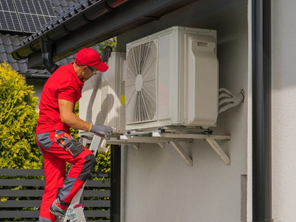 A technician in red uniform works on an outdoor air conditioning unit mounted on the wall of a building, ready to address any HVAC Replacement Questions.