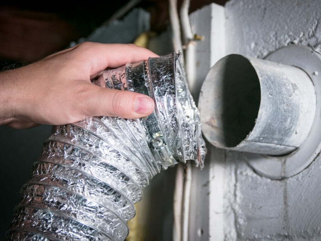 A hand holds a flexible aluminum dryer vent hose near a circular metal duct opening on a wall, an area where mold in HVAC systems can sometimes develop if not properly maintained.