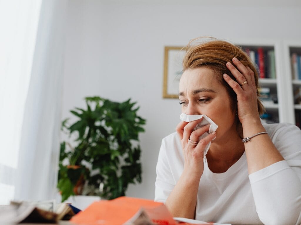 A woman in a white shirt sits at a table holding a tissue to her nose, looking distressed—possibly reacting to mold in HVAC—while papers and a plant are visible in the background.