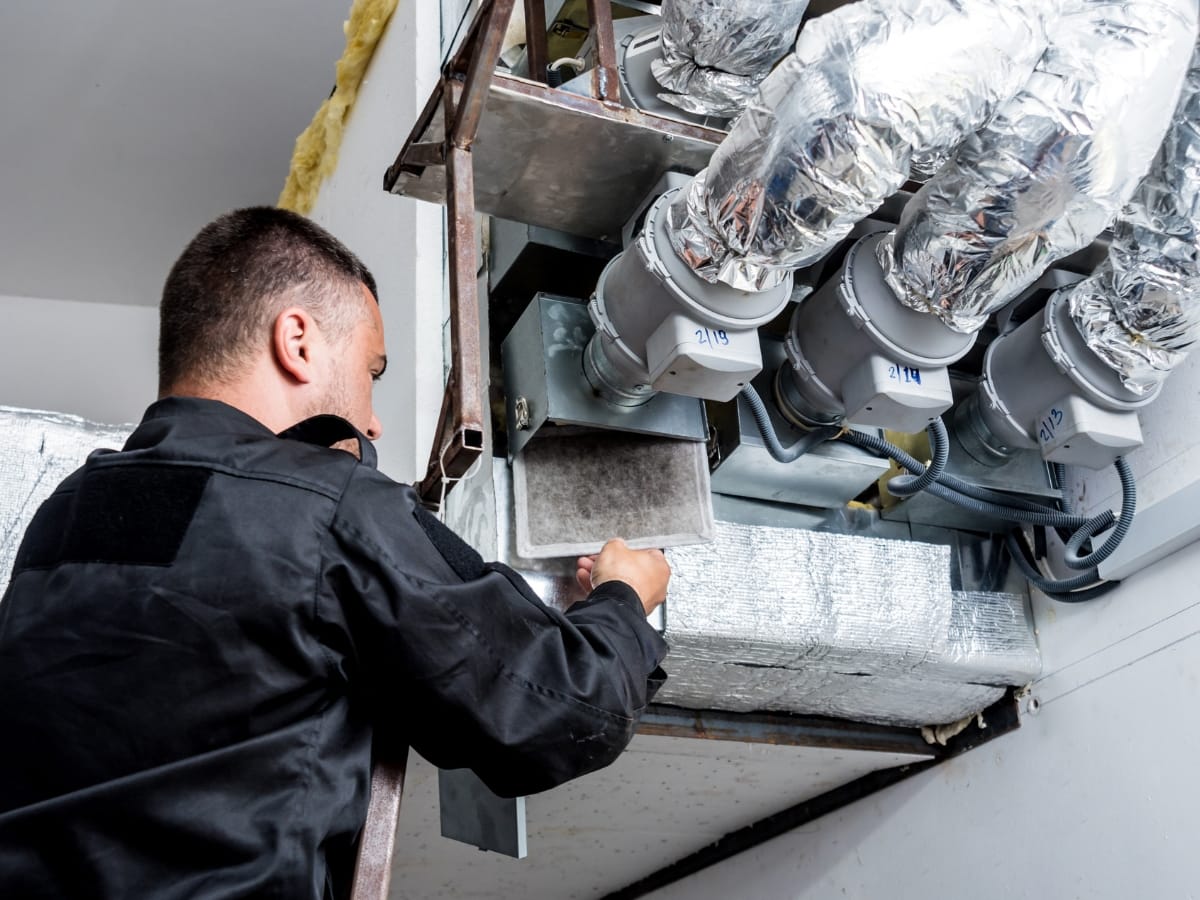 A technician in work clothes replacing or inspecting an air filter in an HVAC duct system with insulated pipes and vents overhead, helping prevent mold in HVAC components.