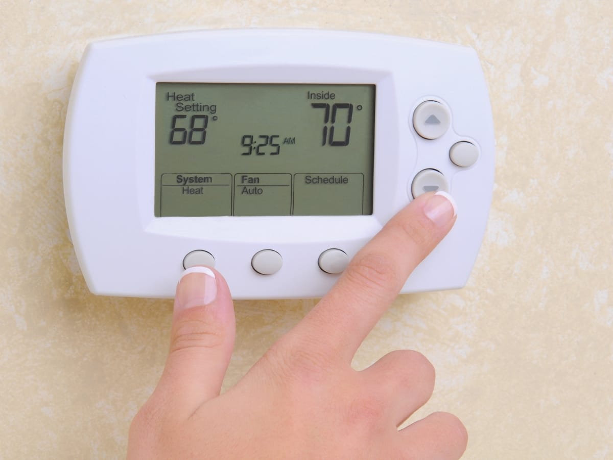 A person adjusting a programmable thermostat set to heat mode, displaying a heat setting of 68°F and an indoor temperature of 70°F.