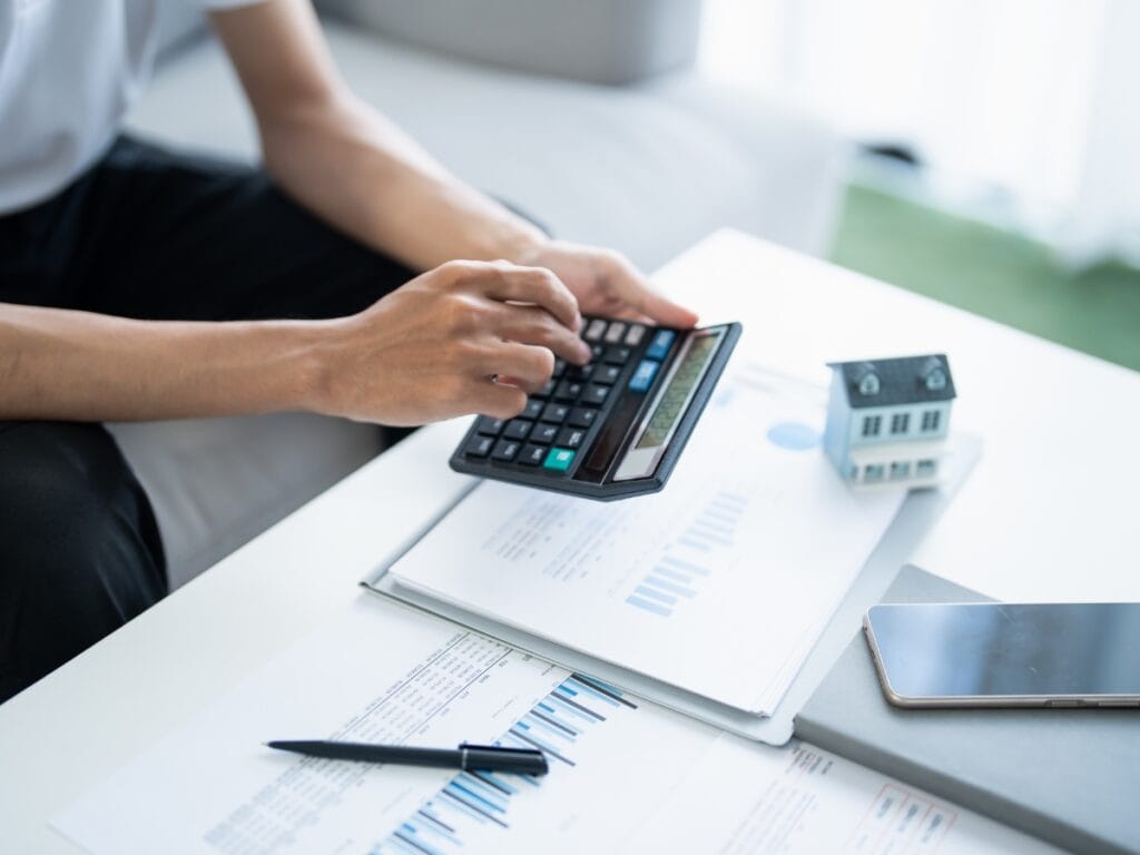 A person uses a calculator on a desk with financial documents, a pen, a model house, a smartphone, and a programmable thermostat—planning home budgets and energy savings efficiently.