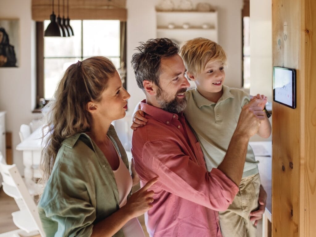 A man holds a boy while adjusting a programmable thermostat on the wall, and a woman stands beside them in a bright, modern kitchen.