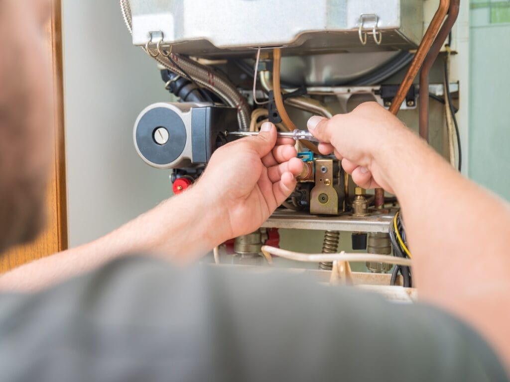 A person uses a wrench to adjust or repair components inside the open panel of a gas boiler, following steps from a furnace maintenance checklist.