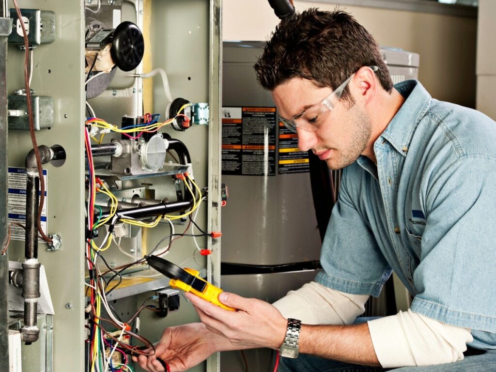 A technician wearing safety glasses uses a multimeter to test electrical connections inside an open furnace unit as part of a thorough furnace maintenance checklist.