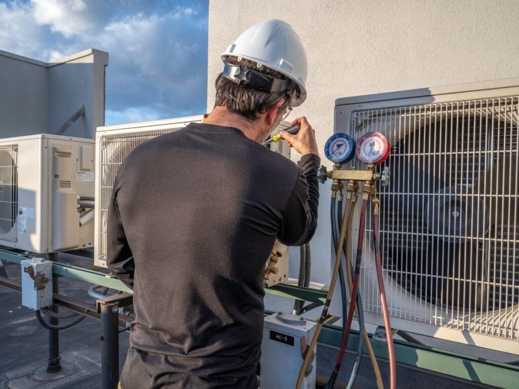 A technician wearing a hard hat uses gauges to service or inspect an air conditioning unit on a rooftop, demonstrating essential end of year HVAC tips for proper system maintenance.