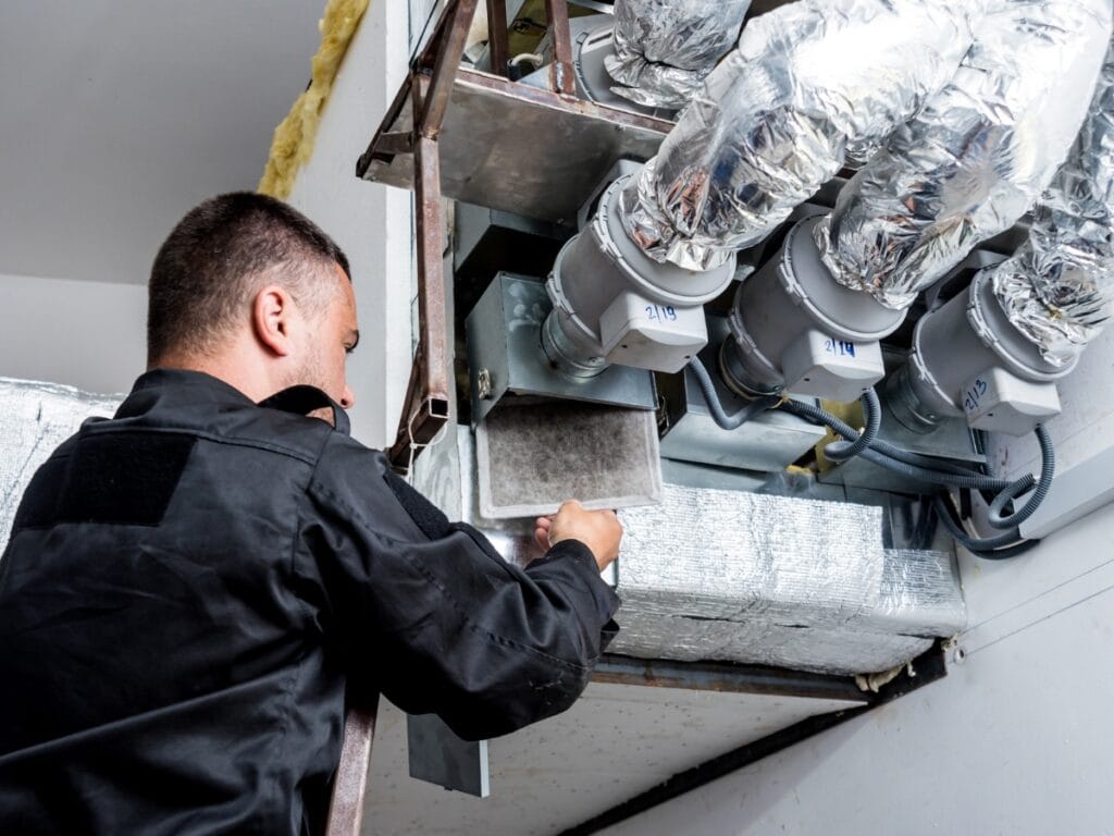 A technician in black clothing replaces an air filter in a large HVAC system with insulated, ducted pipes mounted on the ceiling—perfect example of end of year HVAC tips to keep your system running efficiently.