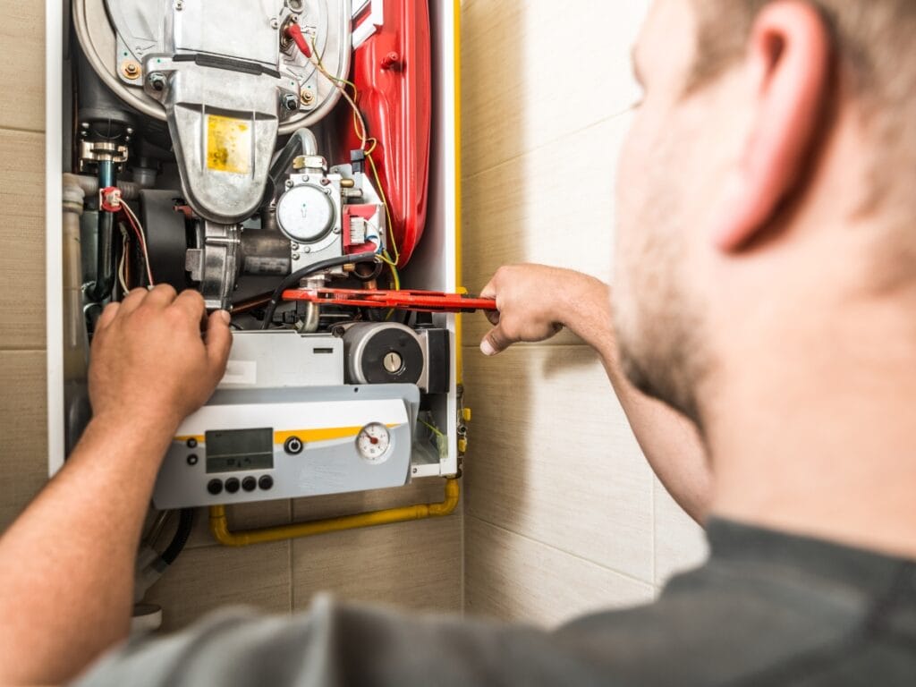 Winterize HVAC Systems: How to Prepare for Freezing Temps 4 A person uses a wrench to adjust components inside an open gas boiler mounted on a wall, helping to winterize HVAC systems for the colder months.