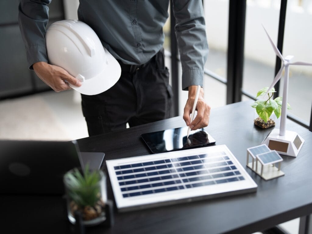 Winterize HVAC Systems: How to Prepare for Freezing Temps 5 Person in business attire holding a white hard hat and using a tablet, with a solar panel, wind turbine model, and laptop on a desk—reviewing plans to winterize HVAC systems for sustainable energy management.