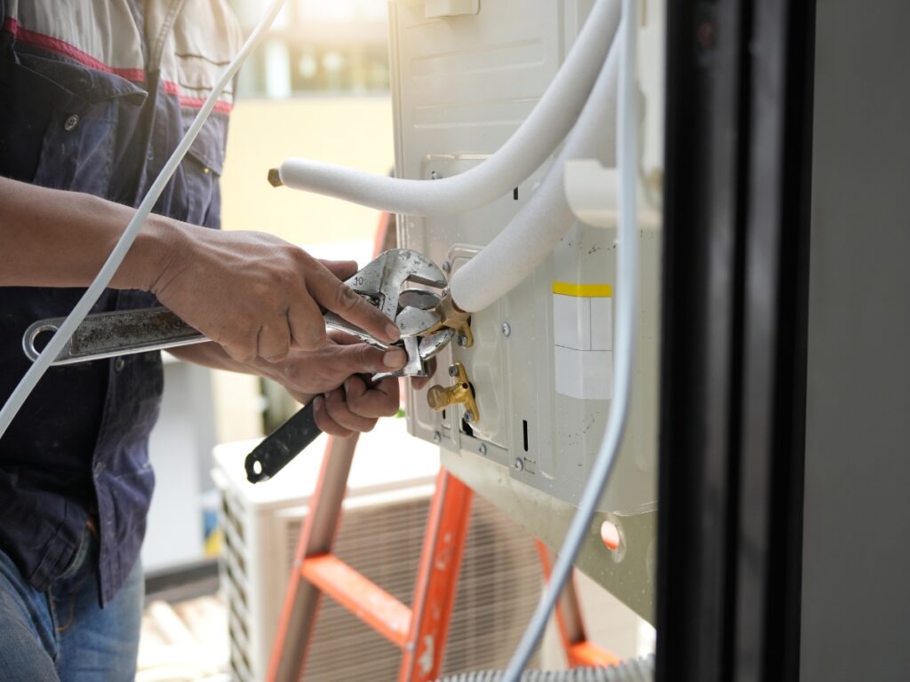 A person uses a wrench to adjust beginner-friendly components of pipes connected to an outdoor air conditioning unit, with a red ladder in the background.