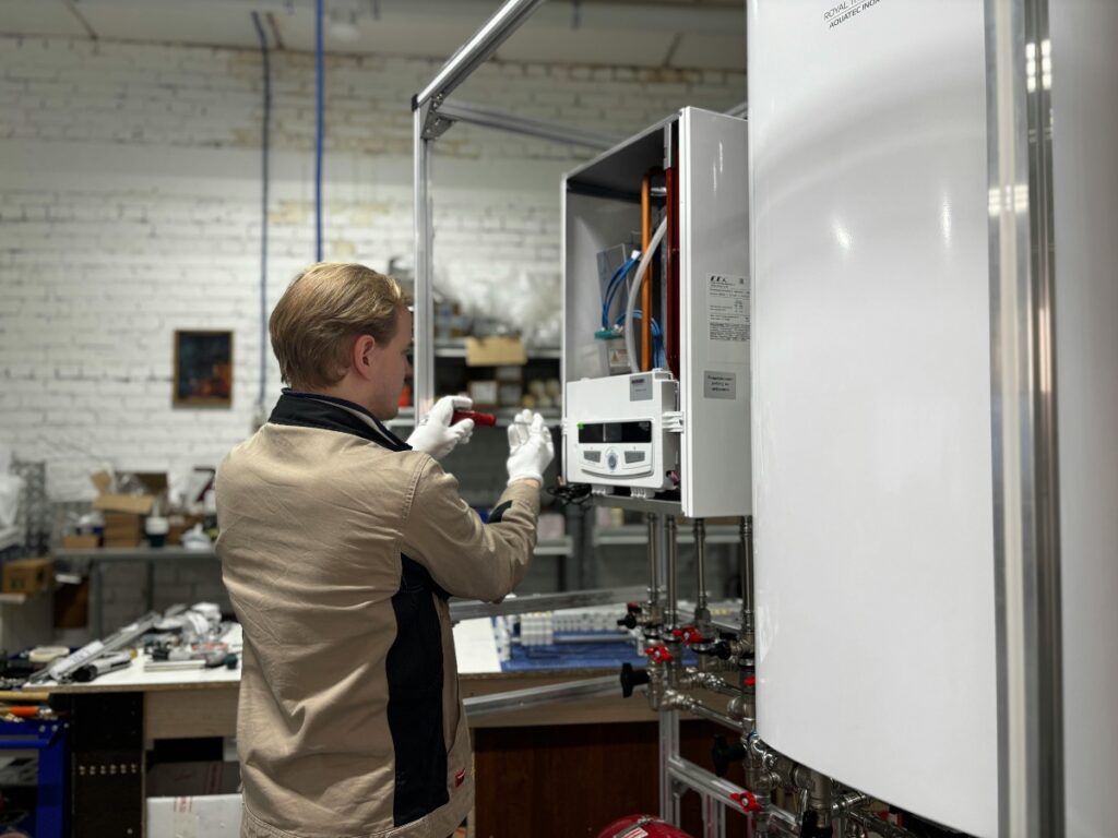 Person wearing gloves and a uniform uses a tool to service or inspect an open wall-mounted boiler, demonstrating key components for beginners in an industrial workshop.