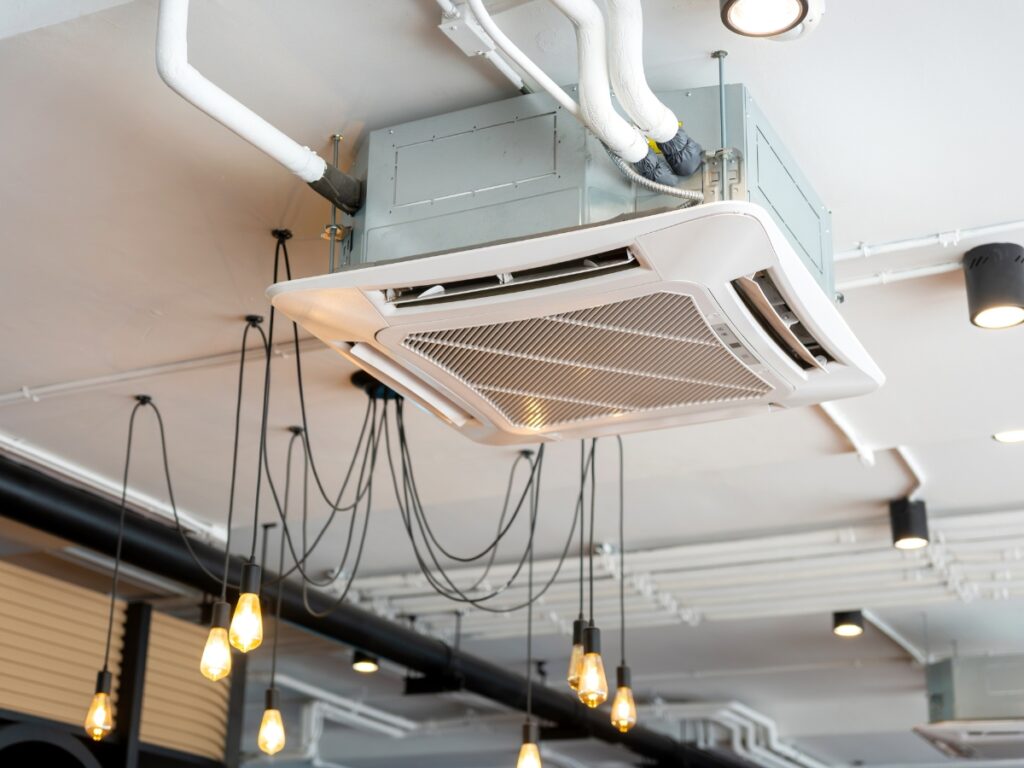 Ceiling-mounted cassette air conditioner unit, ideal for beginners, surrounded by hanging light bulbs and exposed piping on a white ceiling highlighting essential components.