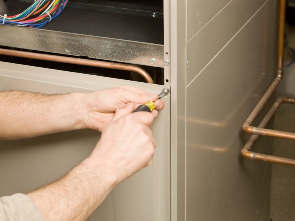 Person using a screwdriver to loosen or tighten a screw on the metal panel of an electric furnace, with wiring and copper pipes visible.