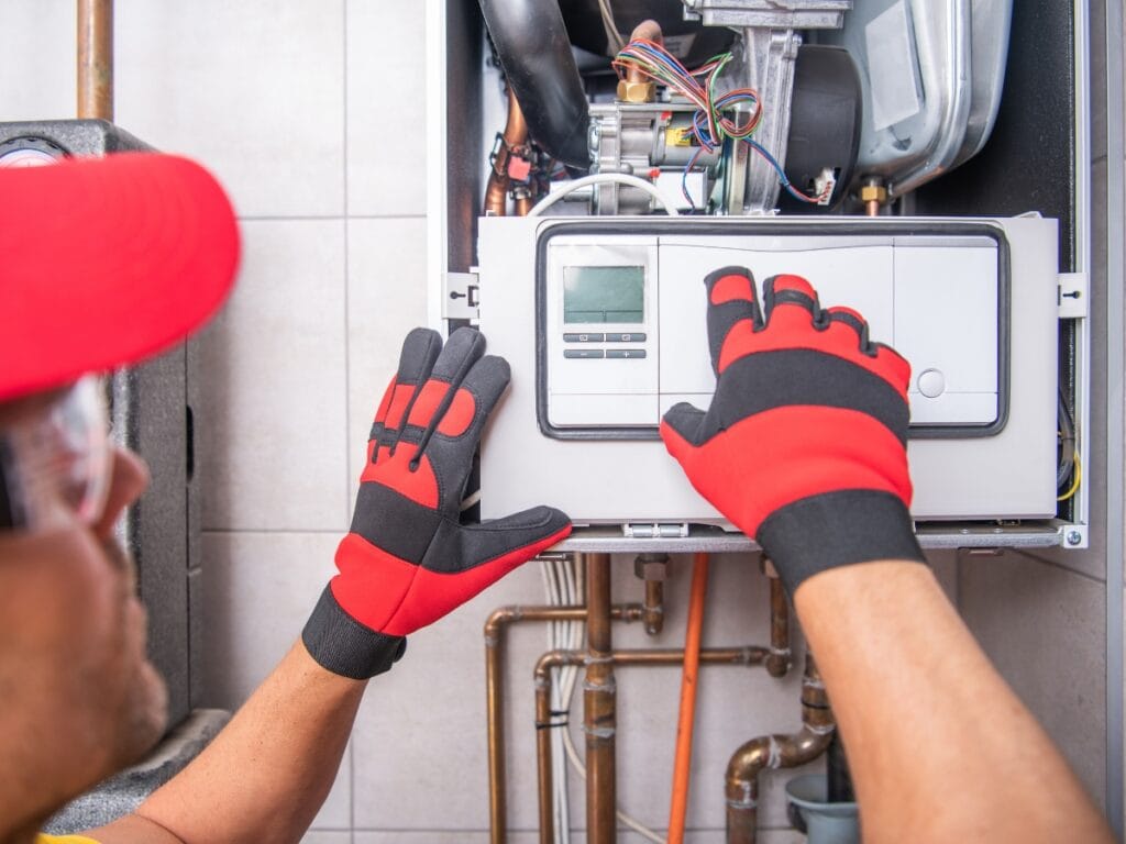 A technician wearing red and black gloves adjusts the controls on a wall-mounted electric furnace or heating system with exposed internal components.