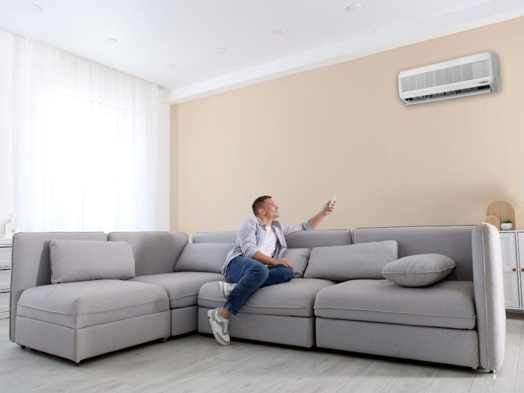 A person sitting on a gray sectional sofa in a modern living room uses a remote to operate a wall-mounted air conditioner, enhancing comfort and indoor air quality with the help of humidifiers.