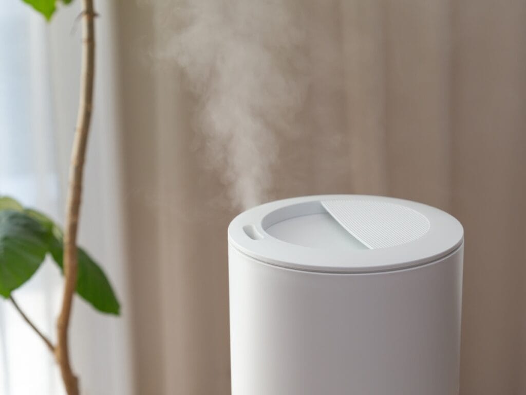 A white indoor air quality humidifier emits visible mist, positioned near a green leafy plant with neutral-colored curtains in the background.