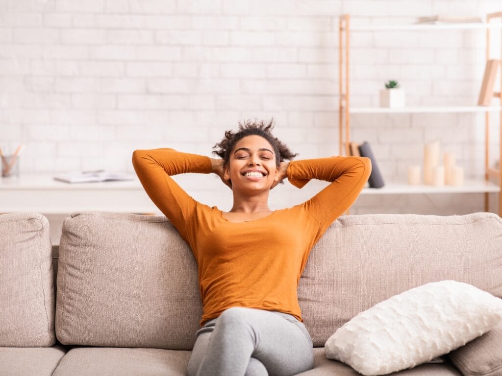 A person in an orange shirt sits on a beige sofa, leaning back with hands behind their head and smiling, enjoying excellent indoor air quality thanks to humidifiers in a bright living room with shelves in the background.