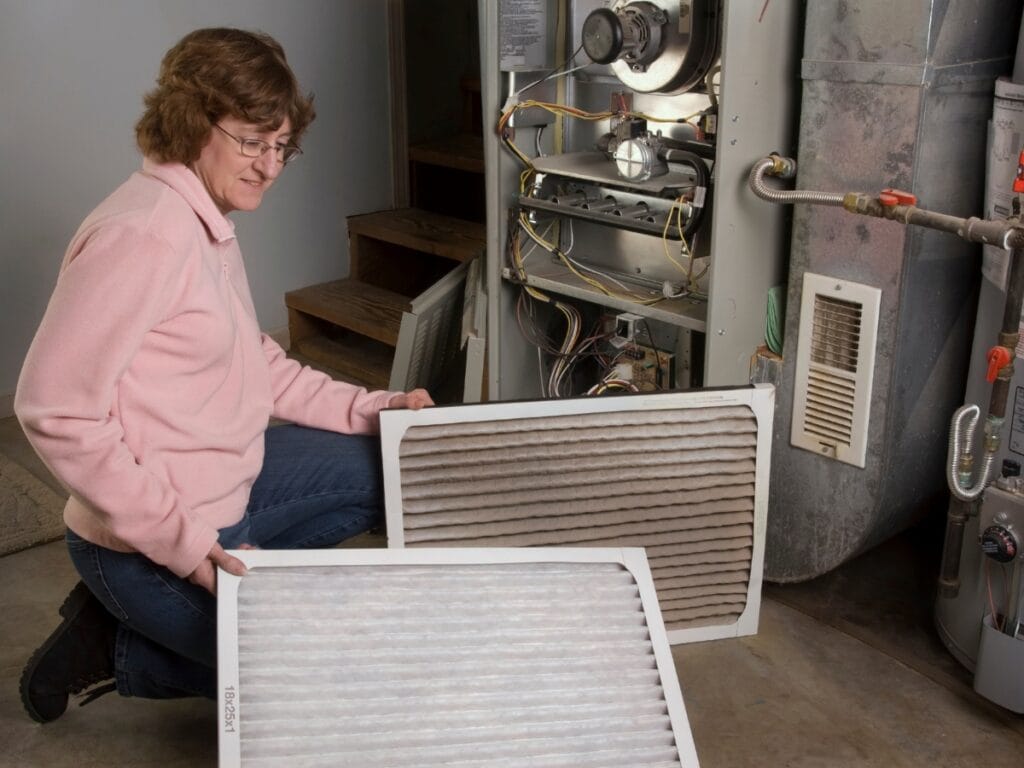 A woman holds a clean and a dirty air filter in front of an open furnace, serving as an air filter guide while demonstrating the difference and maintenance process.
