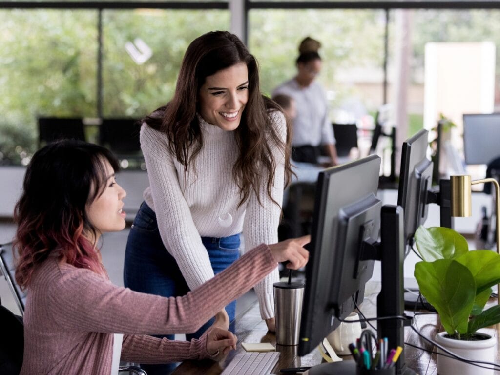 Seasonal Vacation Prep: How to Save Energy Efficiently While Away 3 Two women in an office setting look at a computer monitor together, one seated and pointing at the screen while the other stands and smiles, collaborating on seasonal vacation prep tasks.