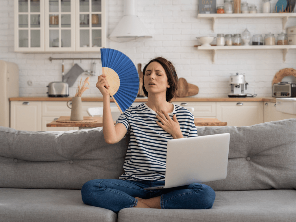 A woman sits cross-legged on a couch with a laptop, fanning herself with a blue fan and appearing overheated in a kitchen setting, wishing for air conditioning.