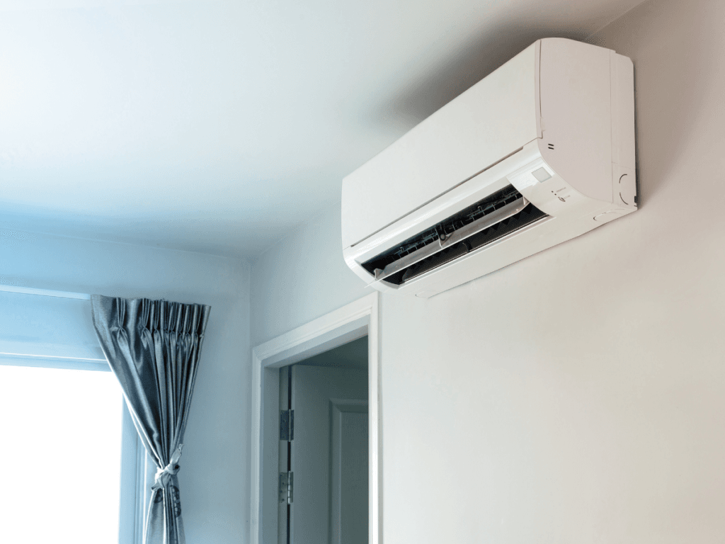 A white wall-mounted air conditioning unit is installed above a doorway in a room with light walls and a window with gray curtains for efficient climate control.