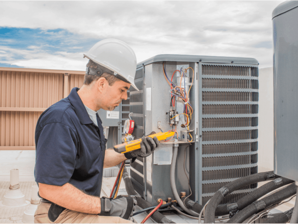 A technician wearing a hard hat and gloves uses a multimeter to inspect and service an outdoor air conditioning unit on a rooftop, ensuring the air conditioning system runs efficiently.