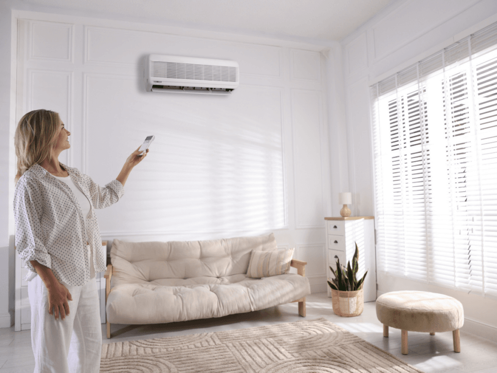A woman in a bright living room uses a remote control to operate the air conditioning unit mounted on the wall above a sofa.
