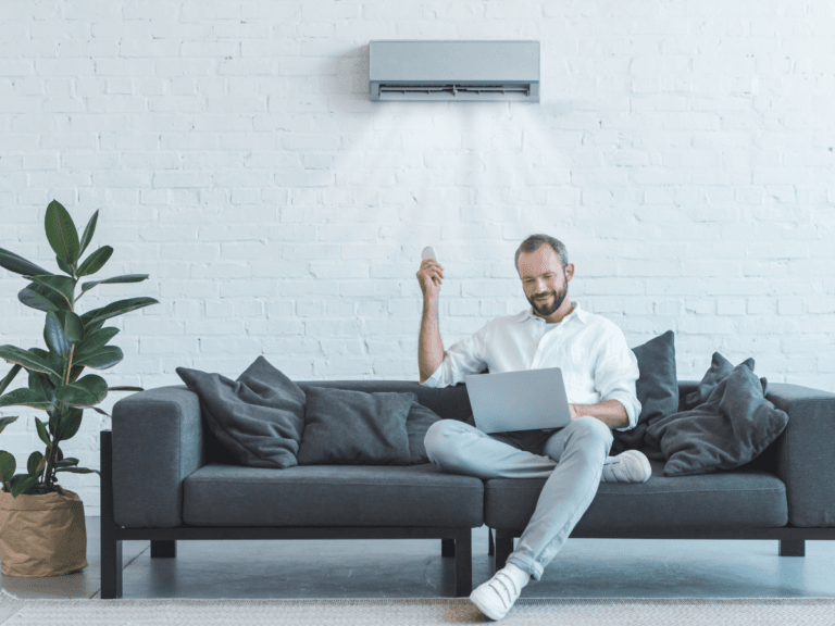 A man sits on a dark gray sofa using a laptop and holding a remote, enjoying the comfort of air conditioning from the unit operating above him on a white brick wall.