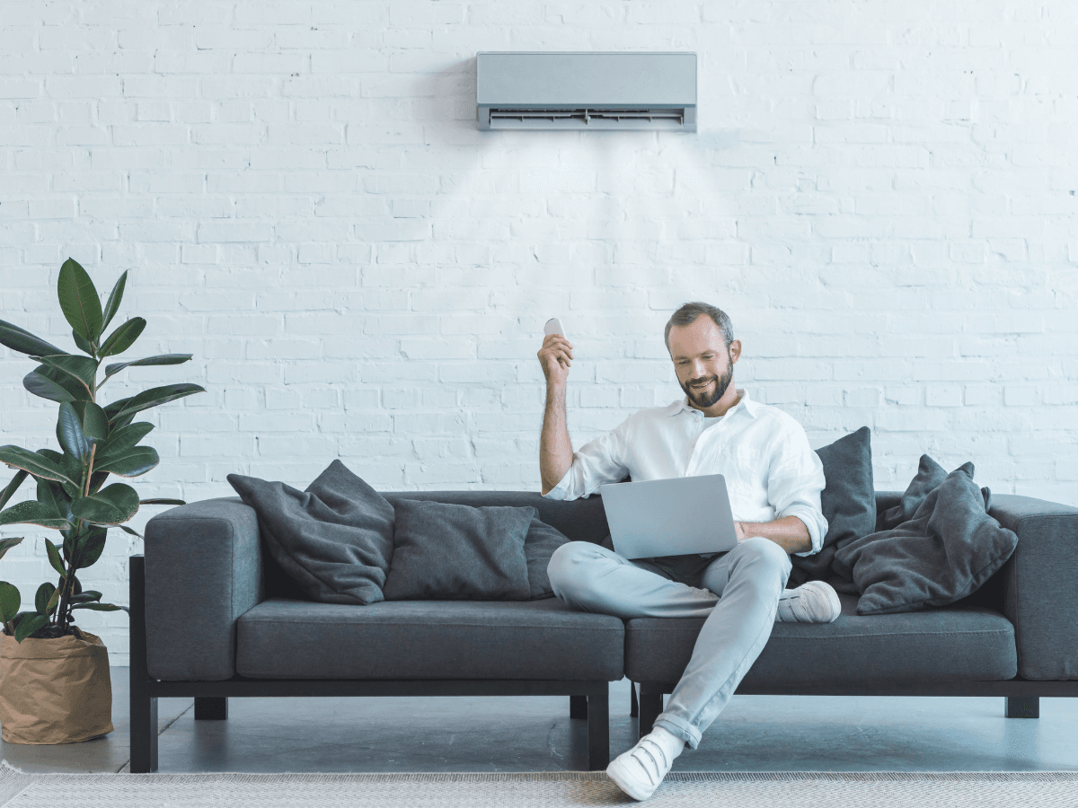 A man sits on a dark gray sofa using a laptop and holding a remote, enjoying the comfort of air conditioning from the unit operating above him on a white brick wall.