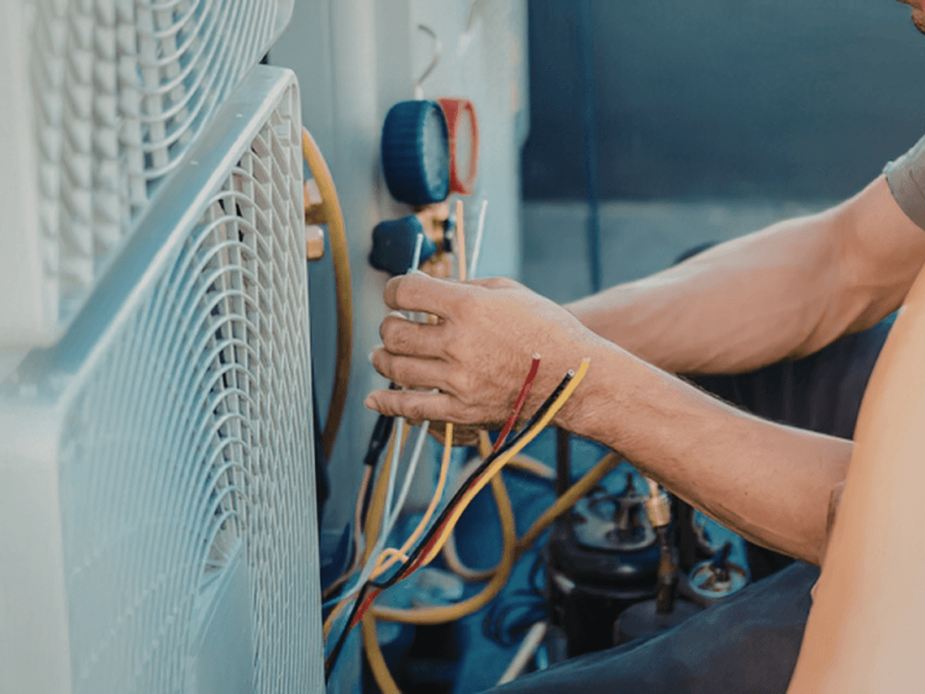 A technician prepares for Summer AC Prep by inspecting an HVAC unit, holding multiple colored electrical wires near gauges and equipment.