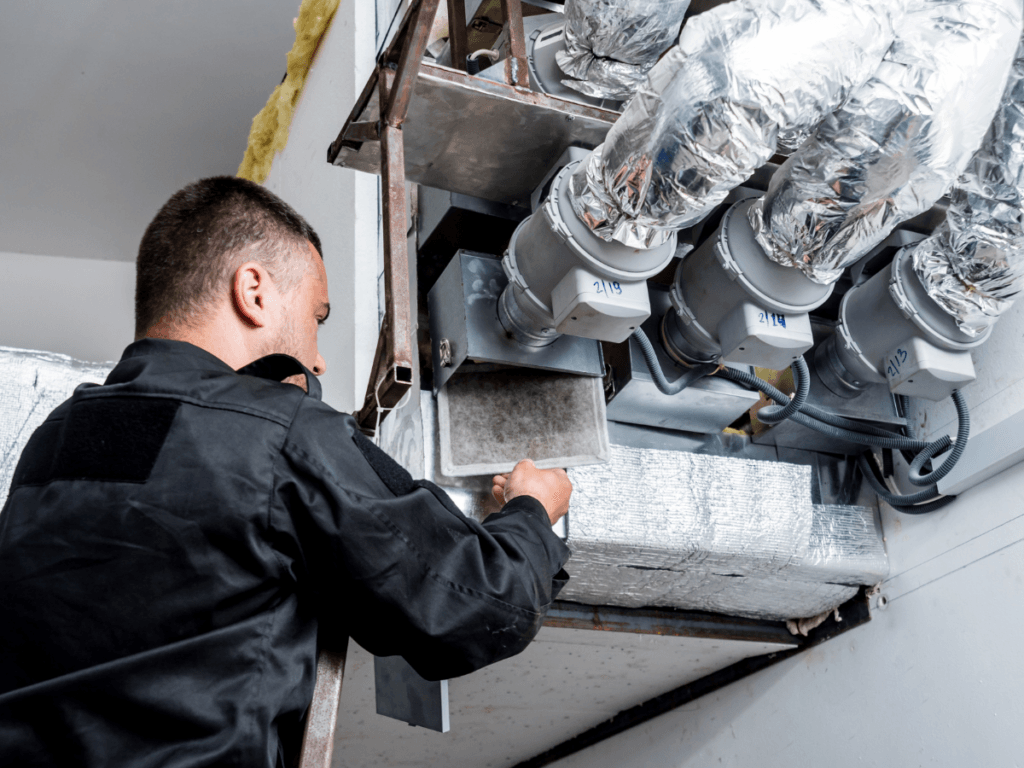 A maintenance worker in uniform inspects or replaces a filter in a commercial HVAC ventilation system with multiple insulated ducts as part of essential Summer AC Prep.