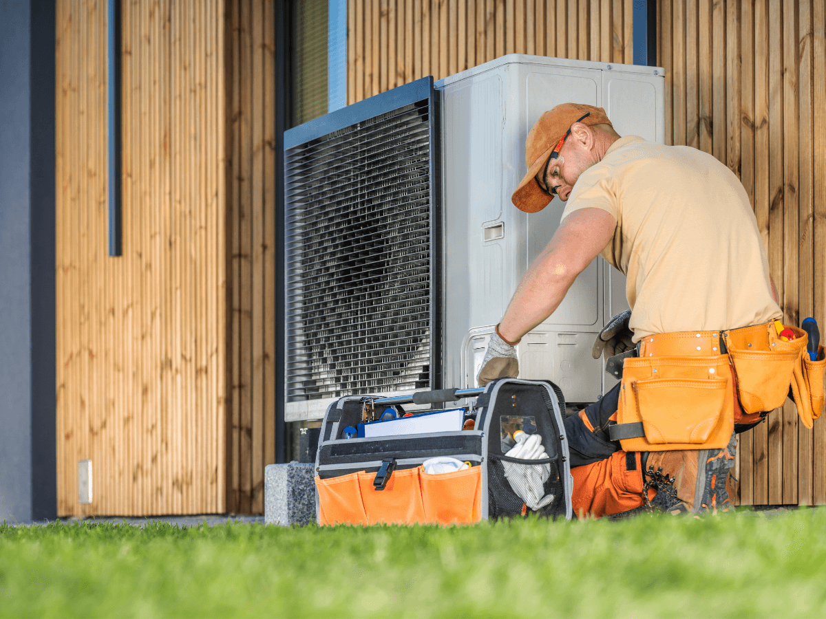 A technician in work attire repairs an outdoor air conditioning unit next to a building, with a tool bag and equipment on the grass nearby, ensuring Summer AC Prep is done for reliable cooling all season long.