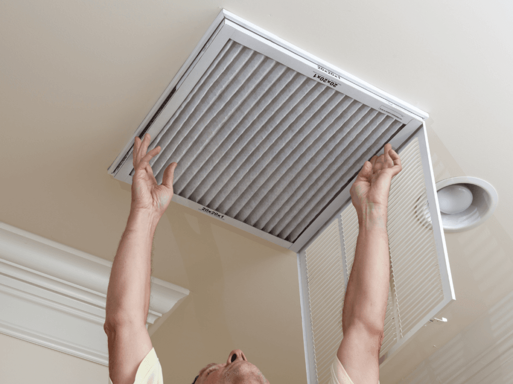 A person installs or replaces an air filter in a ceiling vent, with arms raised and the vent cover open, helping to improve health by reducing pet dander and other airborne particles.