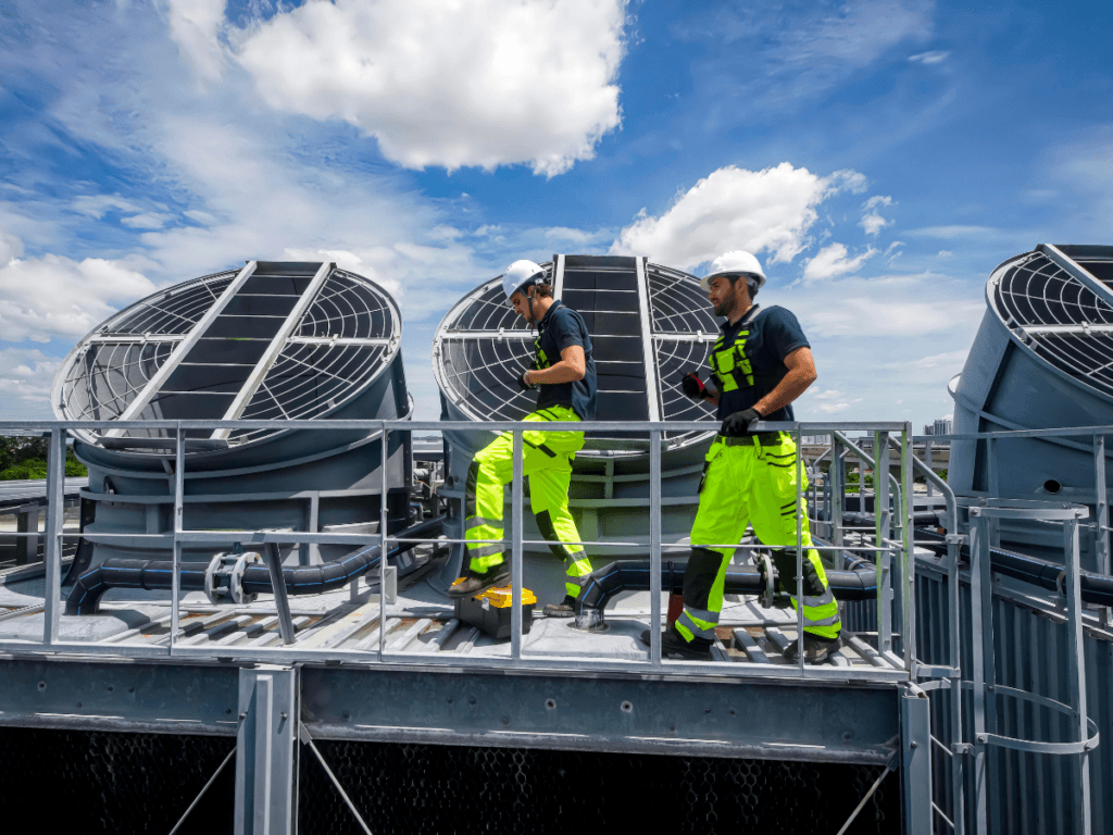Two workers in high-visibility clothing inspect large industrial cooling fans on a rooftop under a partly cloudy sky, preparing for peak seasonal summer demand.