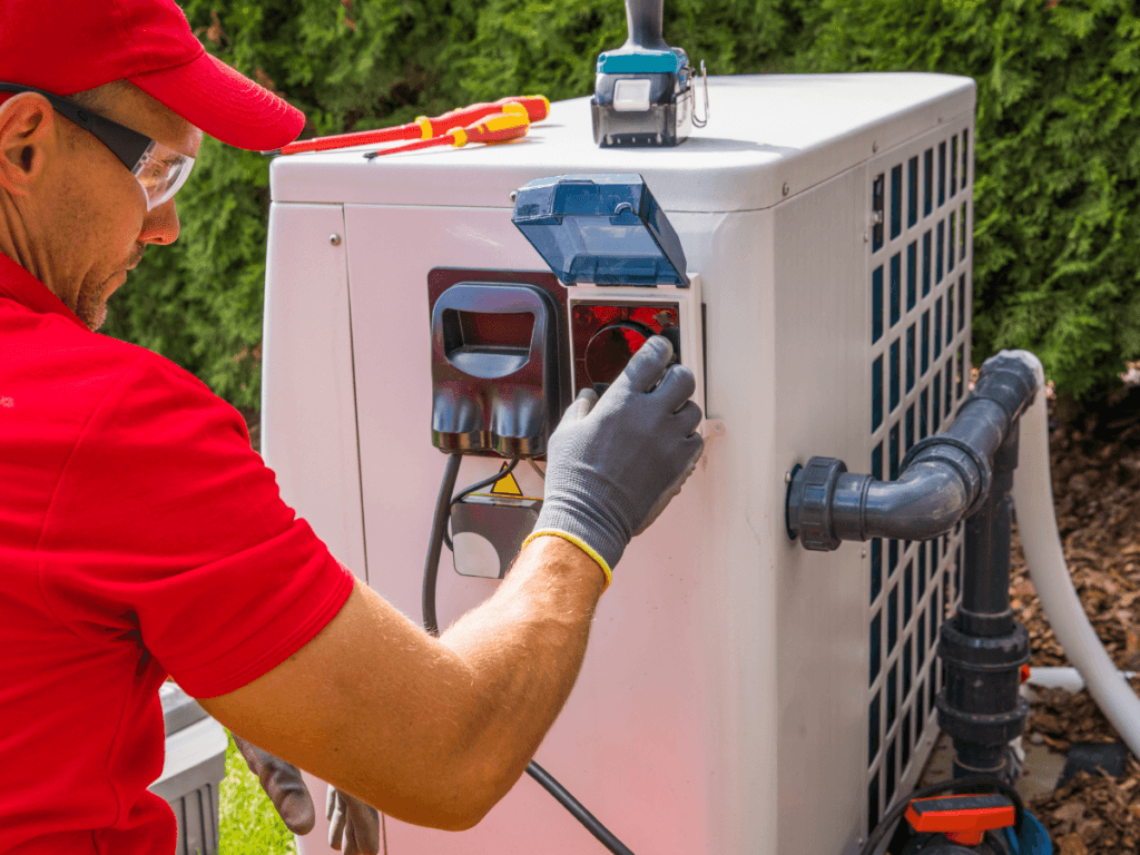Technician in red uniform adjusts settings on an outdoor heat pump unit using a control panel during seasonal summer maintenance, with tools placed on top of the equipment.